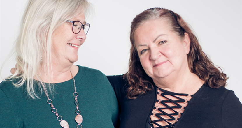 Två kvinnor håller armen om varandra. Kvinnan till vänster har långt grått hår och en mörkgrön tröja. Kvinnan till höger har långt kastanjefärgat hår och en svart tröja. Foto.