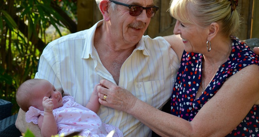 Ett sittande äldre par, där mannen håller ett spädbarn i famnen. Foto.
