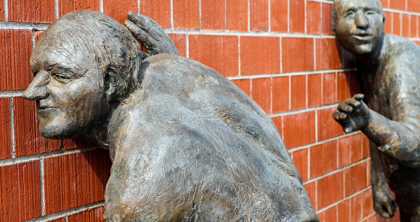 En skulptur med två personer som lyssnar med örat mot en tegelvägg.
