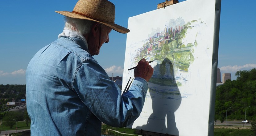 En äldre man står ute i naturen och målar en tavla. 