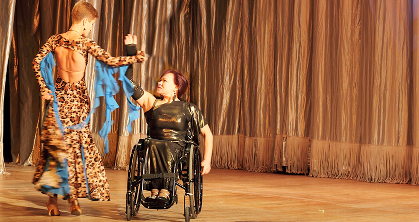 Två kvinnor dansar tango, den ena i rullstol. Båda är vackert klädda och dansar på en scen.
