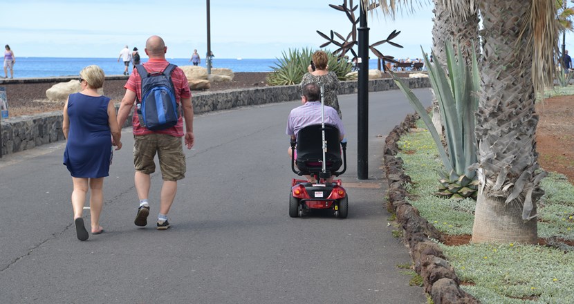 Två personer gående, en i rullstol. Lättklädda på en gata med palmer. Foto.
