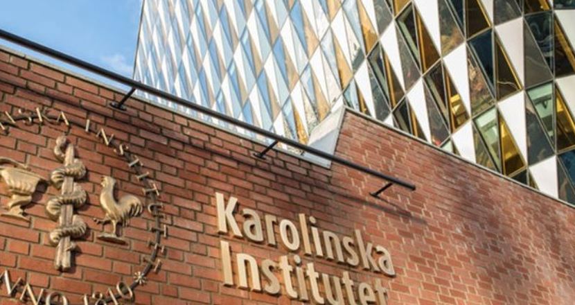 En tegelvägg med texten Karolinska Institutet i förgrunden och en glasfasad med trekanter i bakgrunden. Foto.