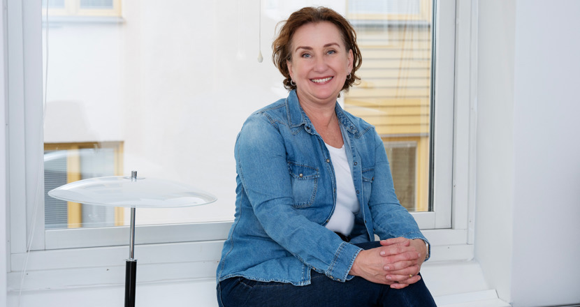 En kvinna i blå jeansskjorta sitter i en fönstersmyg och har ett ljust hus bakom sig och till höger framför sig har hon en vit lampa. Foto.