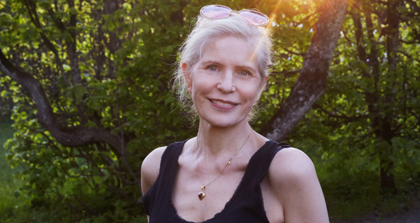 En blond kvinna klädd i ett svart linne med grönska i bakgrunden. Foto.