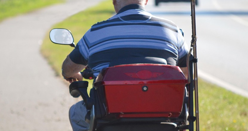 En äldre man på en röd permobil åker ifrån oss på en cykelväg bredvid en väg där en bil och en buss syns i fjärran. Foto.